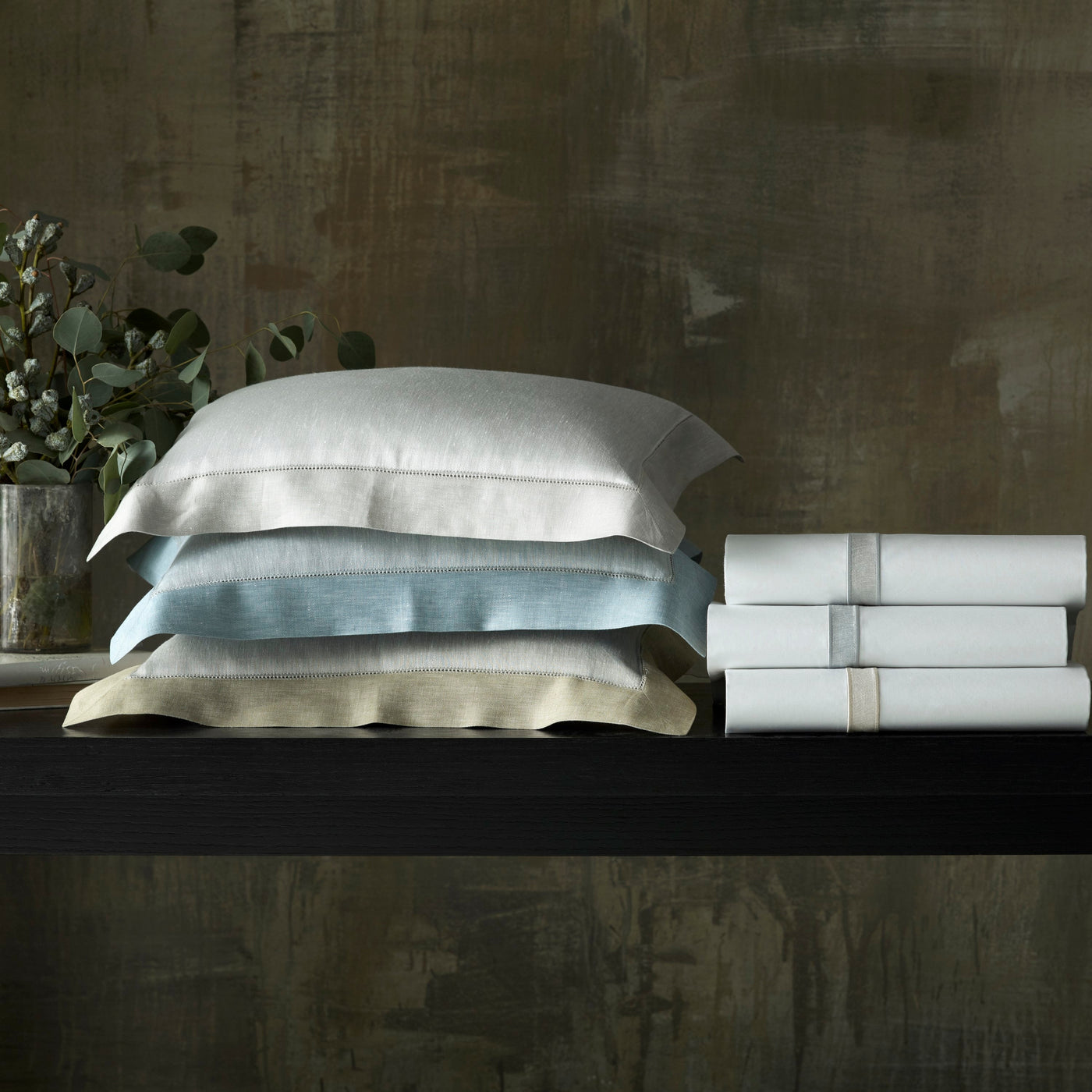 Stacked white, sky blue, and gold linen pillowcases beside folded white sheet sets on dark shelf.