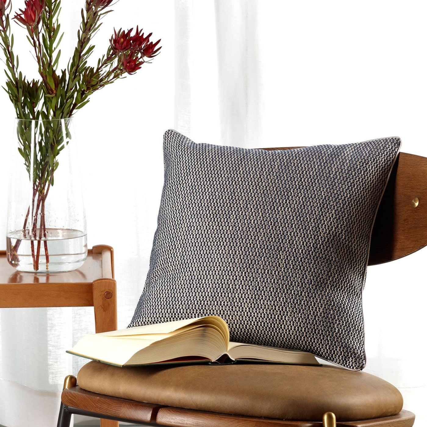 An Amalia Home Collection Jasmim Decorative Pillow with a dobby design rests on a wooden chair with a leather seat, beside an open book; a vase of flowers sits on a nearby table by the window.