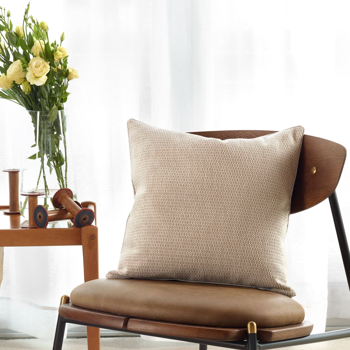 An Amalia Home Collection Jasmim Decorative Pillow with a dobby design sits on a brown leather and wood chair by a table adorned with yellow flowers and wooden toys, in front of sheer white curtains. Made in Portugal.