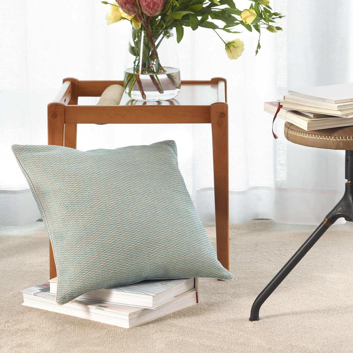 An Amalia Home Collection Jasmim Decorative Pillow with a dobby design sits on stacked books beside a wooden side table topped with a glass vase of flowers, placed on a beige carpeted floor.