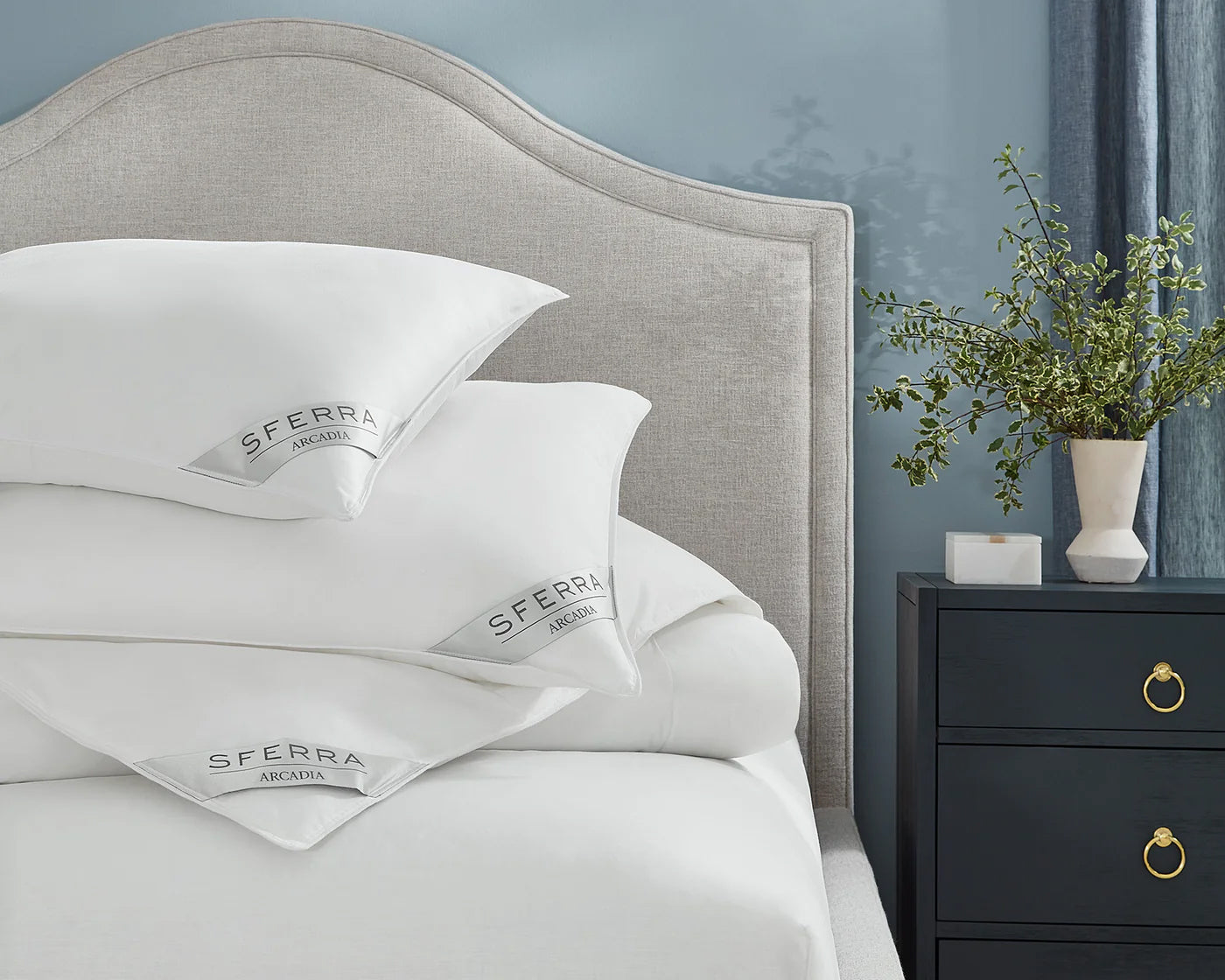 A neatly made bed features SFERRA Arcadia pillows crafted from hypoallergenic microfiber, with stacked cushions, a light upholstered headboard, and a dark nightstand holding a vase of greenery.