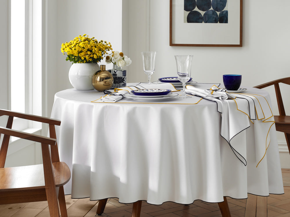 White Home Treasures Arlo round tablecloth styled with navy-trimmed napkins and yellow floral centerpiece.