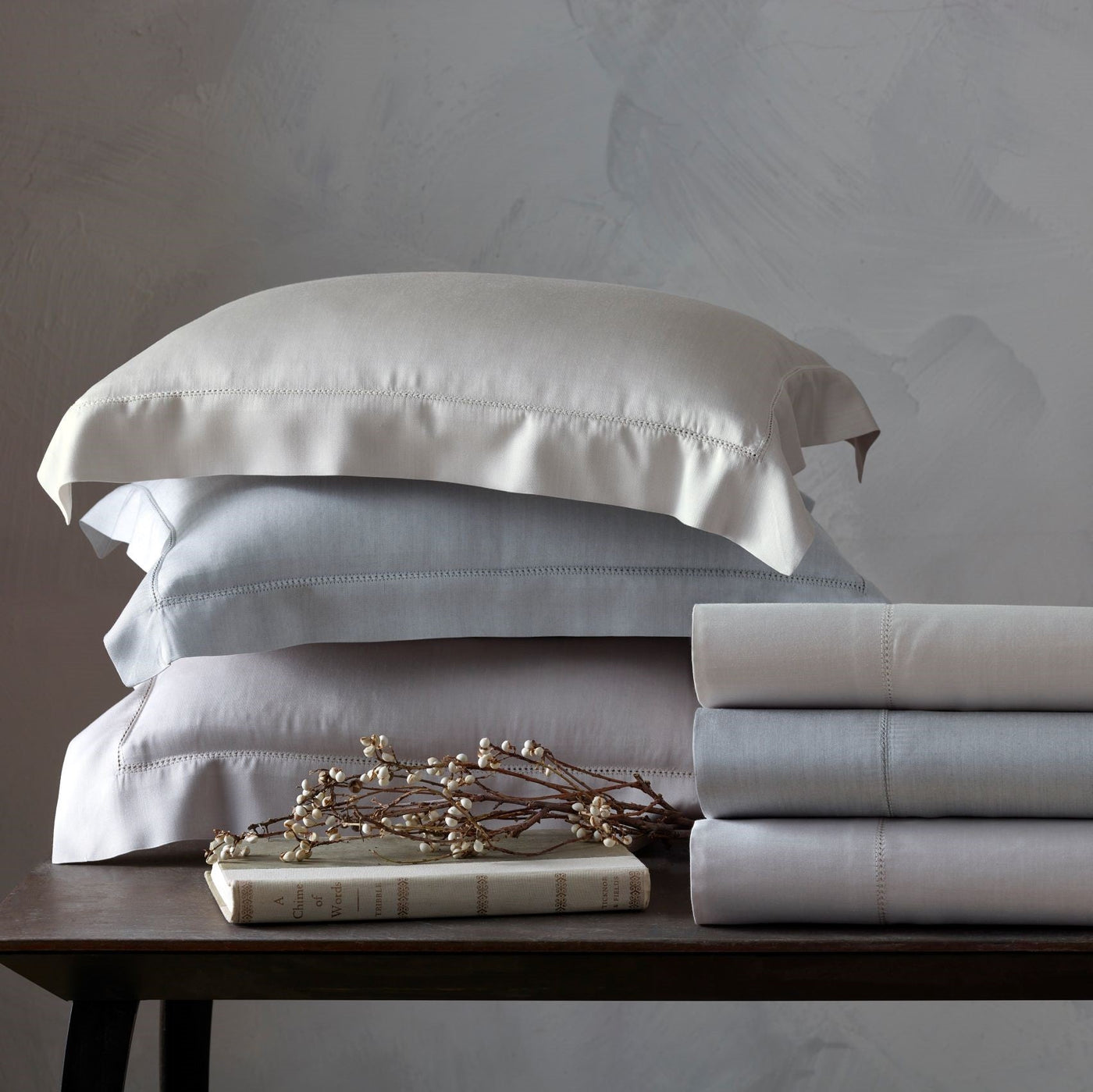 Home Treasures Atwood folded sheets and stacked pillow shams in ivory and silver on wood table.