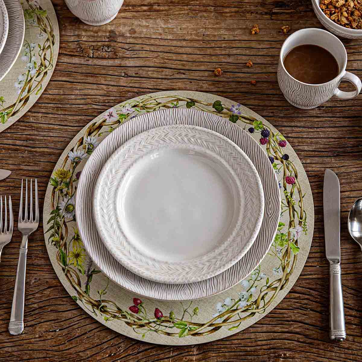 Overhead view of floral botanical placemat layered with white embossed dinnerware on rustic wood table.