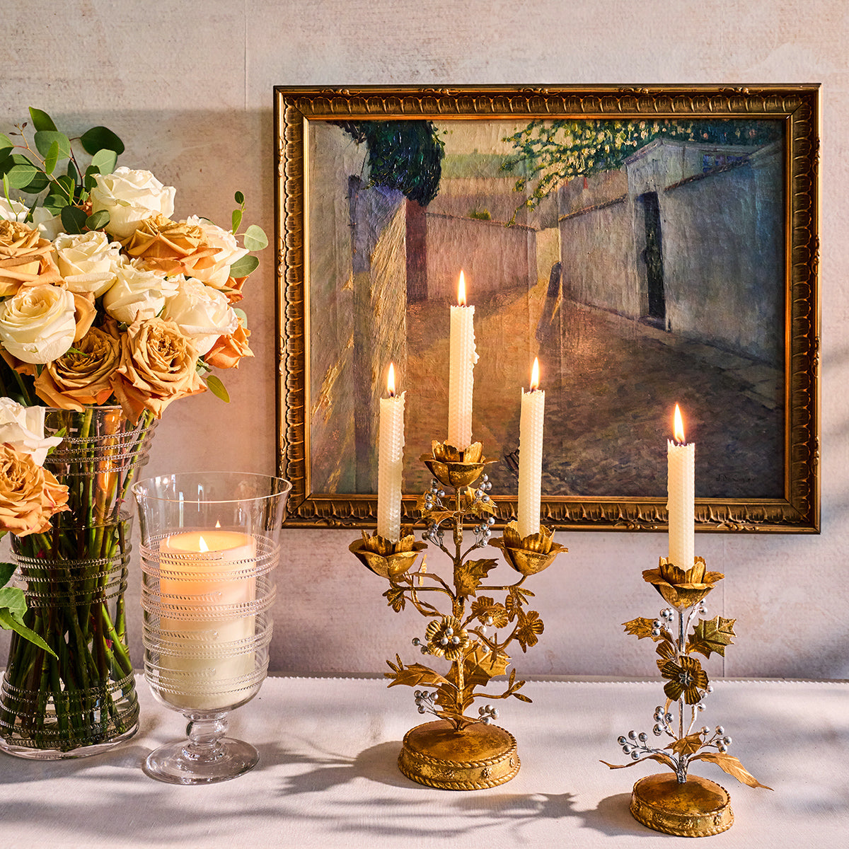 Gold holly candlestick and candelabra with lit tapers on a sideboard beside peach roses