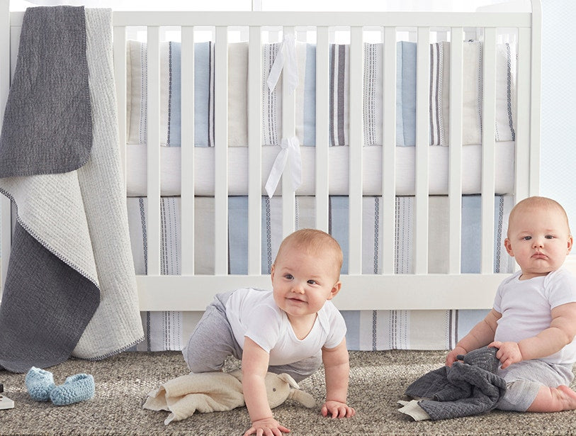 Close-up of grey Coyuchi Cozy Cotton throw draped on white crib with two babies in nursery.