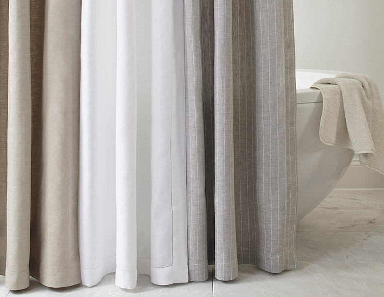 Three linen shower curtains in natural, white, and grey displayed beside a freestanding tub.