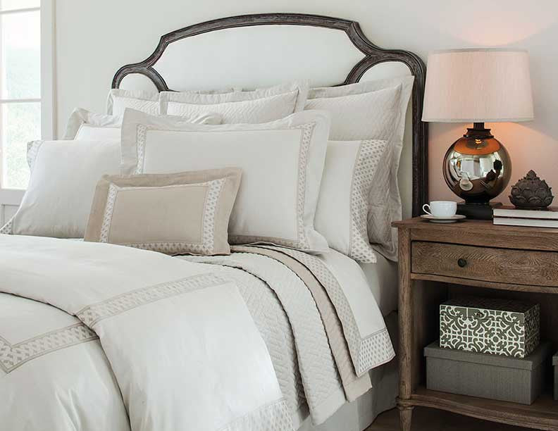 Close-up of ivory bedding with taupe geometric border trim on a wood-framed bed.