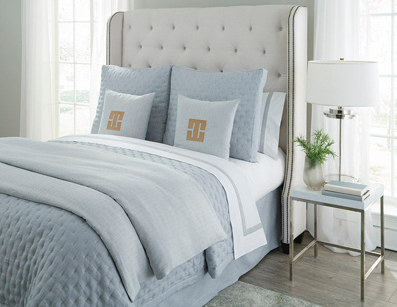 Pale blue quilted yardage draped over a styled bed with monogrammed accent pillows.