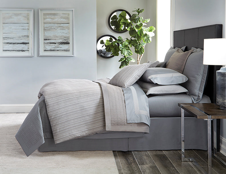 Silver and slate grey layered bedding ensemble styled on a dark upholstered bed.