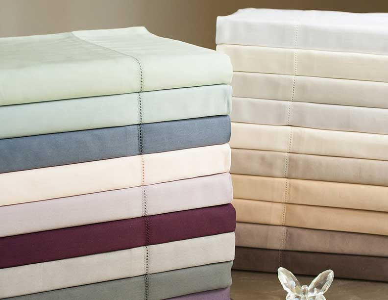 Two stacks of folded Home Treasures Royal sheets in assorted colors with hemstitch detail.
