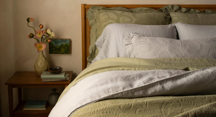 A bed layered in sage and pale grey linen with ruffled jacquard shams and a matching coverlet, beside a nightstand with poppies in a vase.