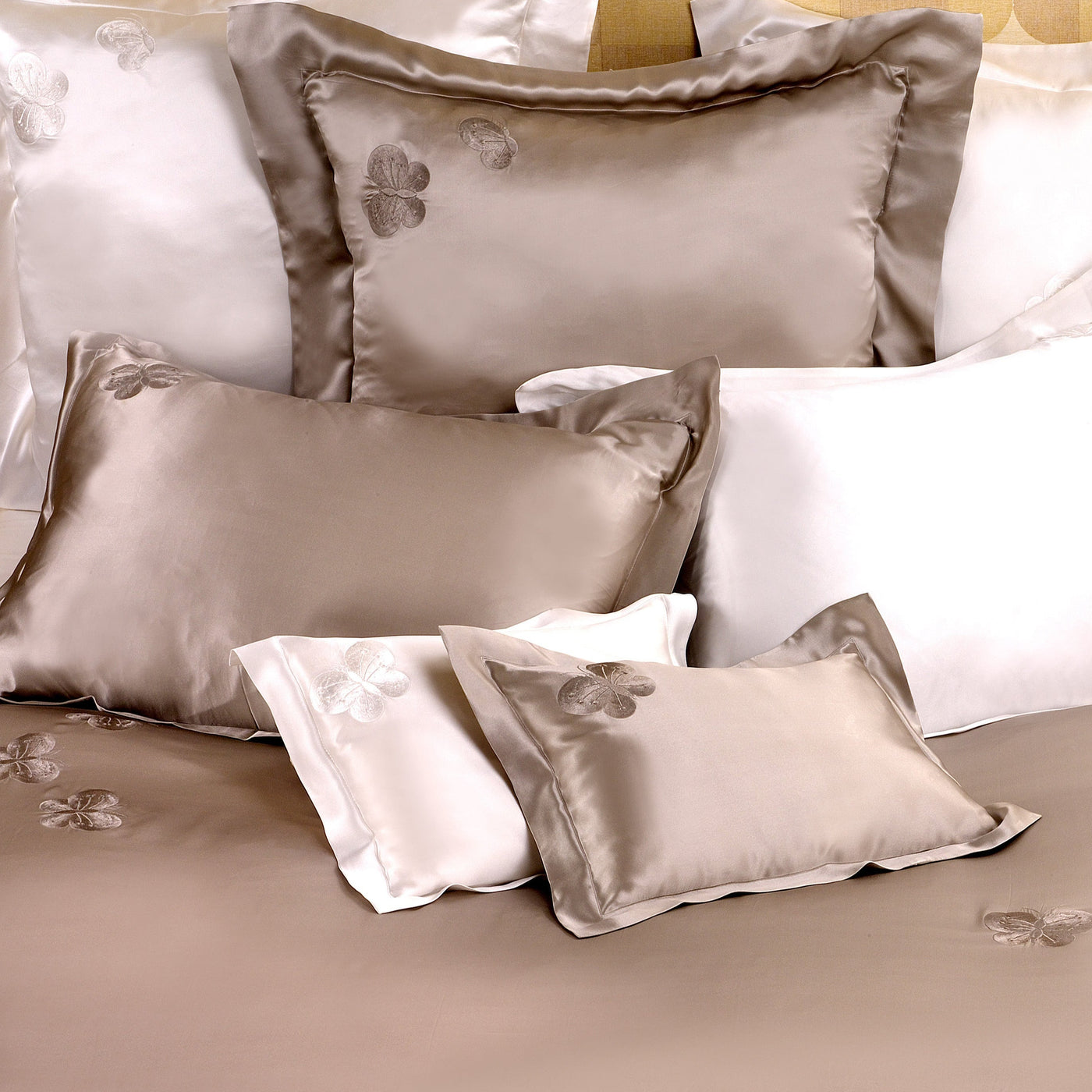 A neatly arranged bed features shiny taupe and white silk charmeuse sheets and Kumi Kookoon Butterfly Silk Shams, some adorned with embroidered butterfly designs.