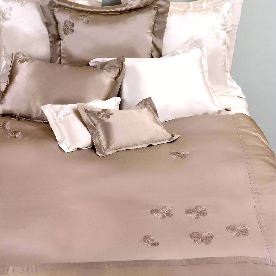 A neatly made bed featuring the Kumi Kookoon Butterfly Silk Duvet Cover, accented with beige and white decorative pillows and a beige comforter embroidered with floral patterns.