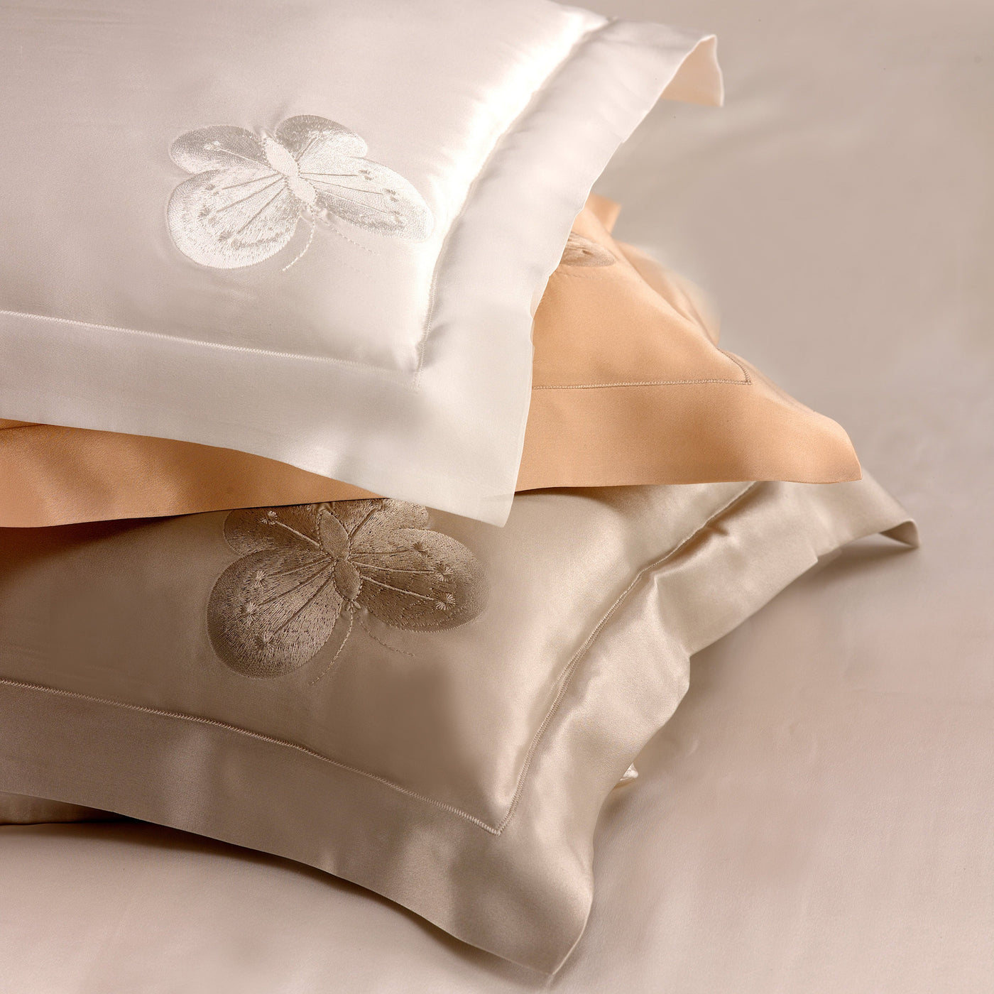 Three Butterfly Silk Shams by Kumi Kookoon—white, beige, and taupe—feature large embroidered flowers on elegant silk charmeuse covers, stacked against a neutral background.
