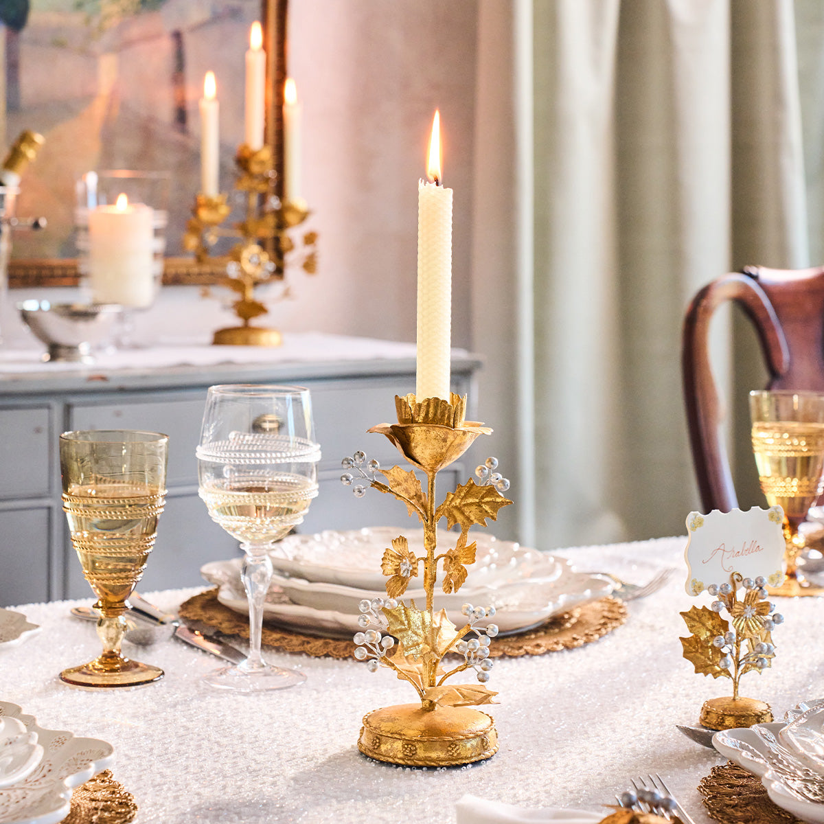 Gold holly candlestick with a lit taper on a table set with amber glasses and white plates