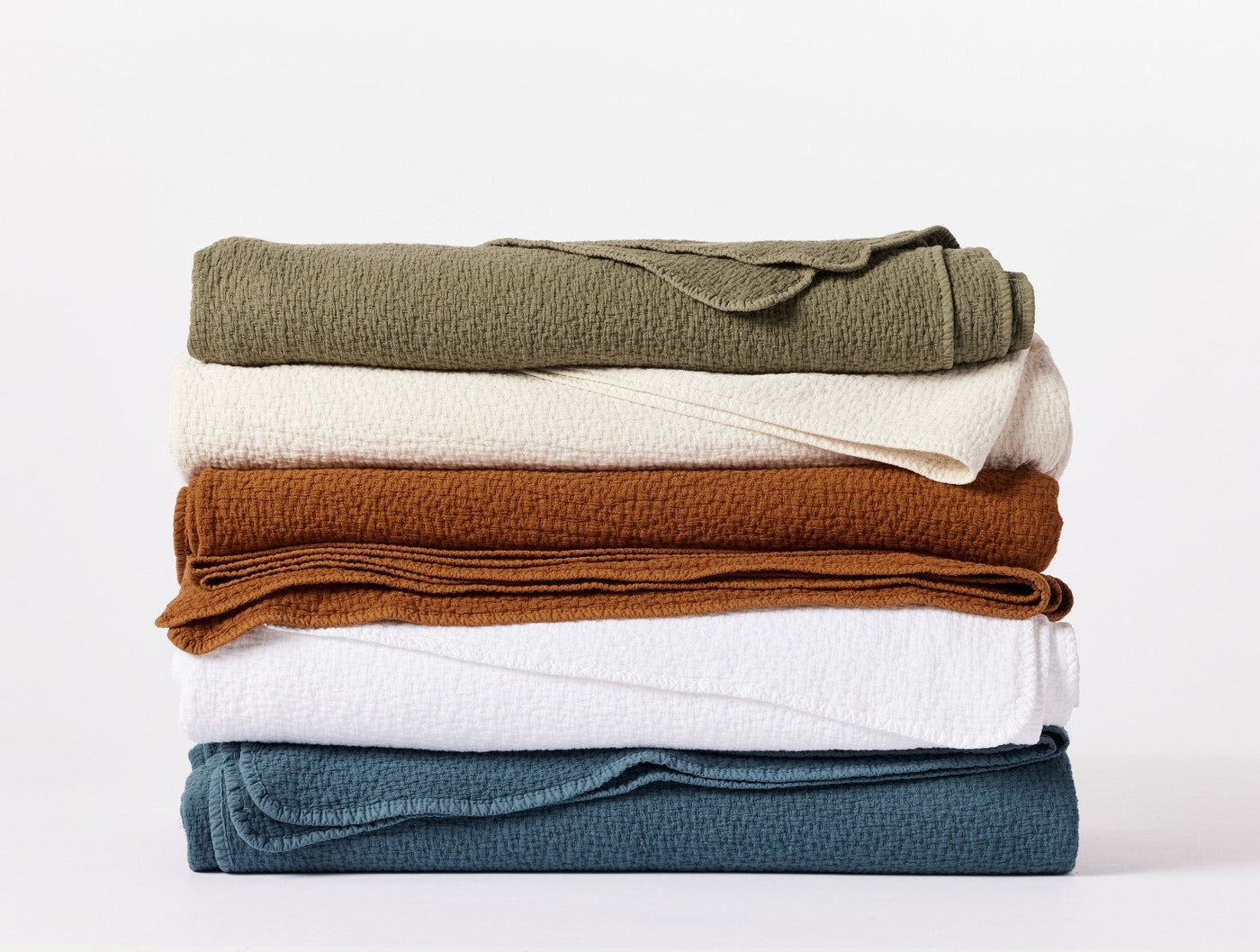 Stack of five folded Coyuchi Cascade Matelasse blankets in olive, ivory, amber, white, and teal.