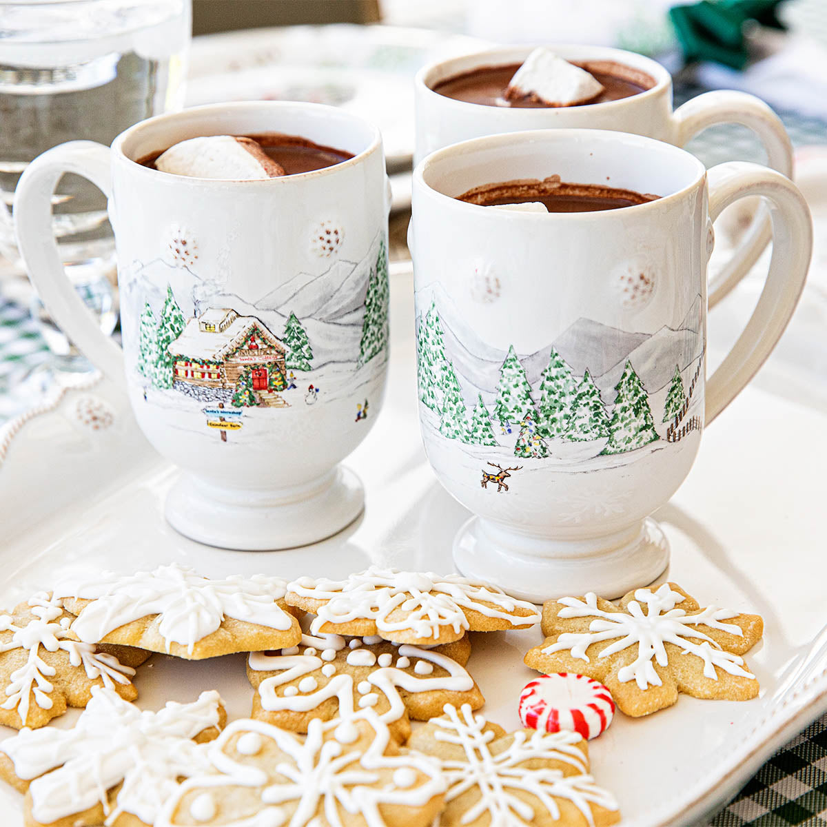 Two North Pole mugs of hot cocoa with marshmallows beside iced snowflake cookies