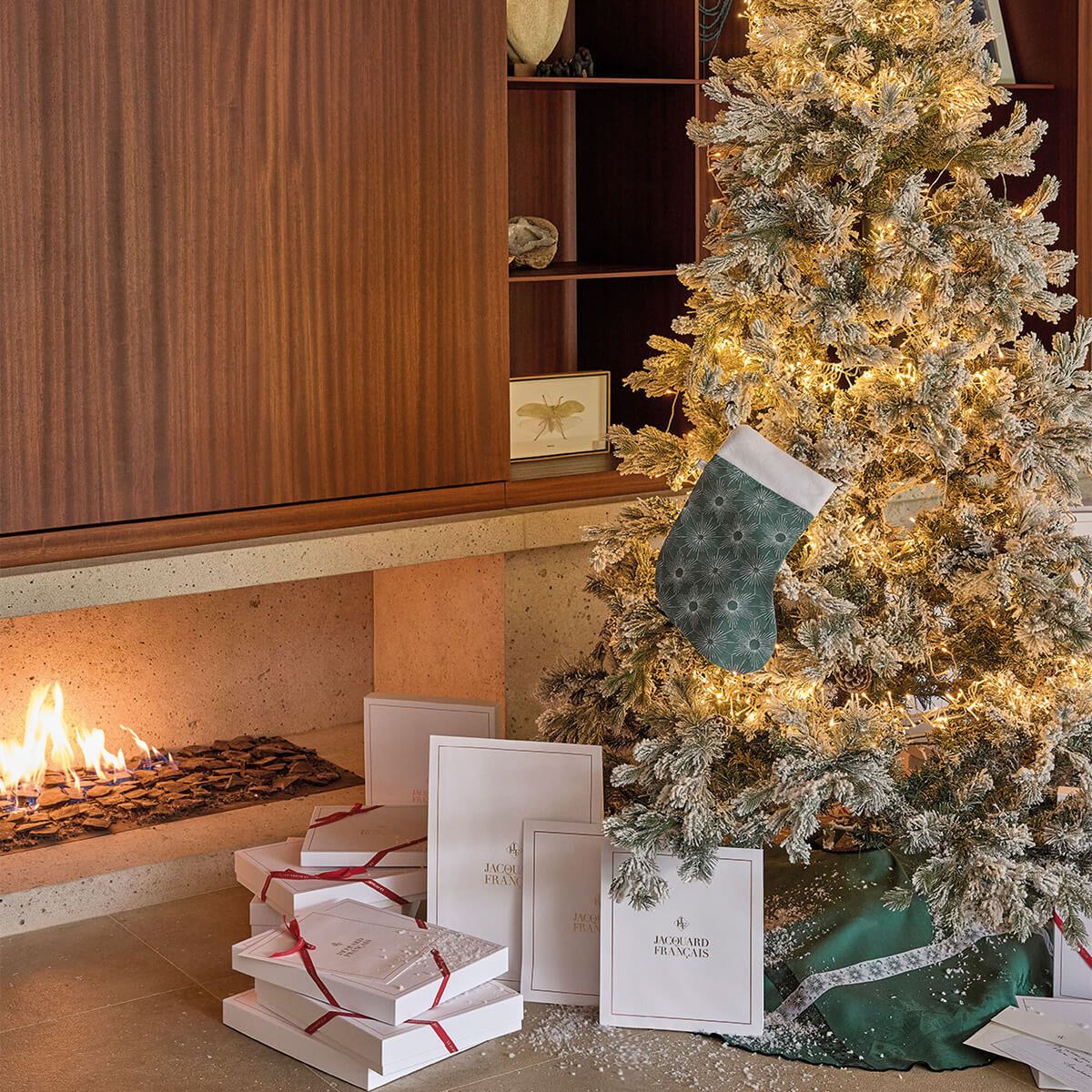 Le Jacquard Français Green Christmas stocking hanging on a flocked tree beside a lit fireplace.