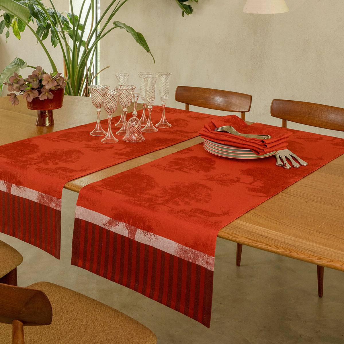 Two red Souveraine linen table runners laid across a wooden dining table set with glasses and napkins