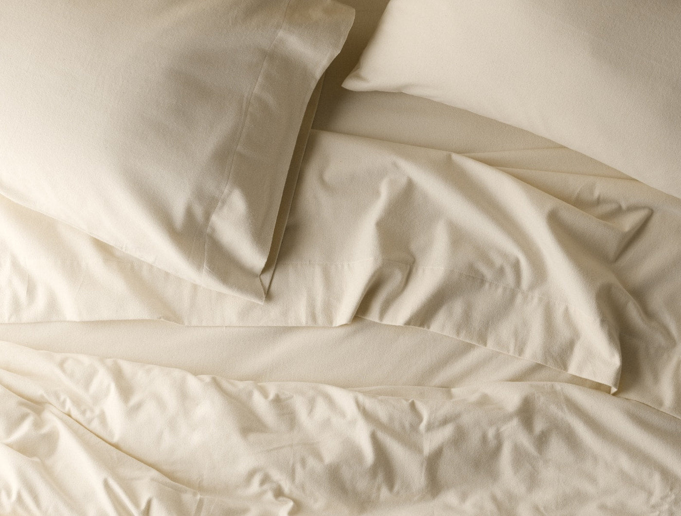 Softly rumpled ivory flannel sheets and pillowcase draped across a bed, overhead view.