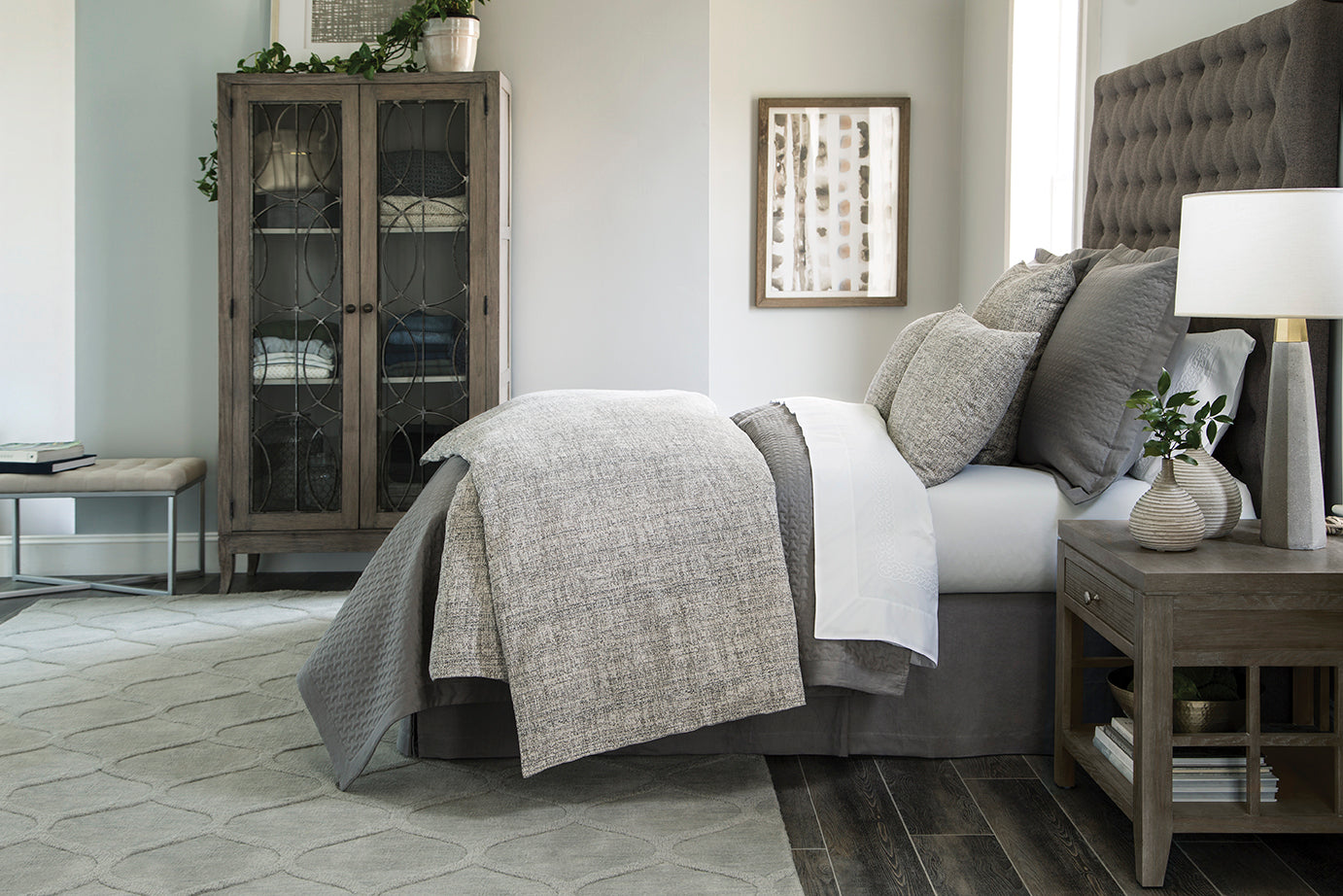 Layered gray bedding with textured quilt on upholstered bed in neutral bedroom setting.