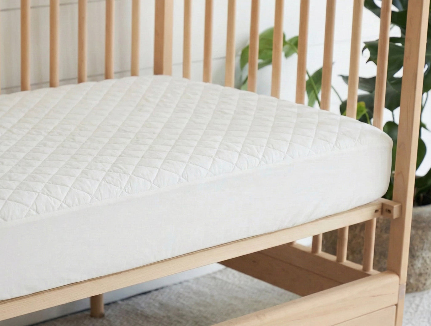 Quilted ivory cotton mattress pad fitted on a natural wood crib, angled view.