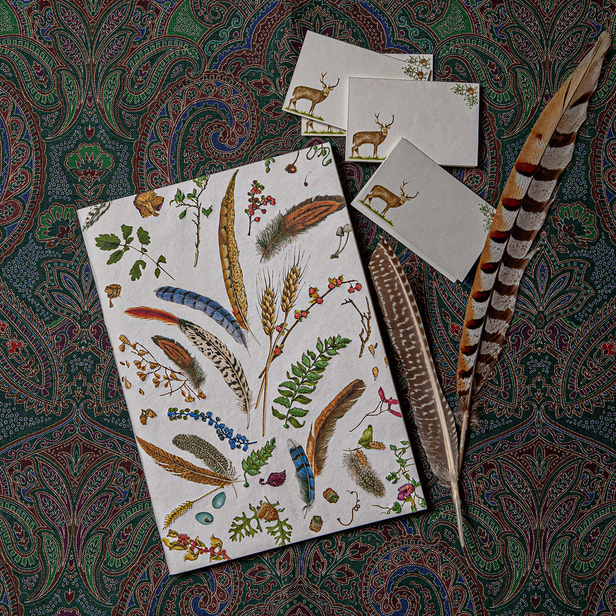 Forest Walk feather-print journal and stag cards on a paisley cloth beside a pheasant feather