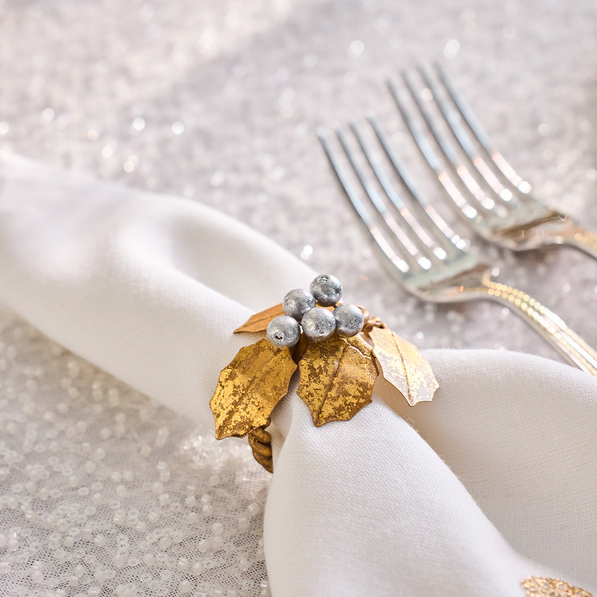 Close-up of a gold holly napkin ring with silver berries on a white napkin beside forks