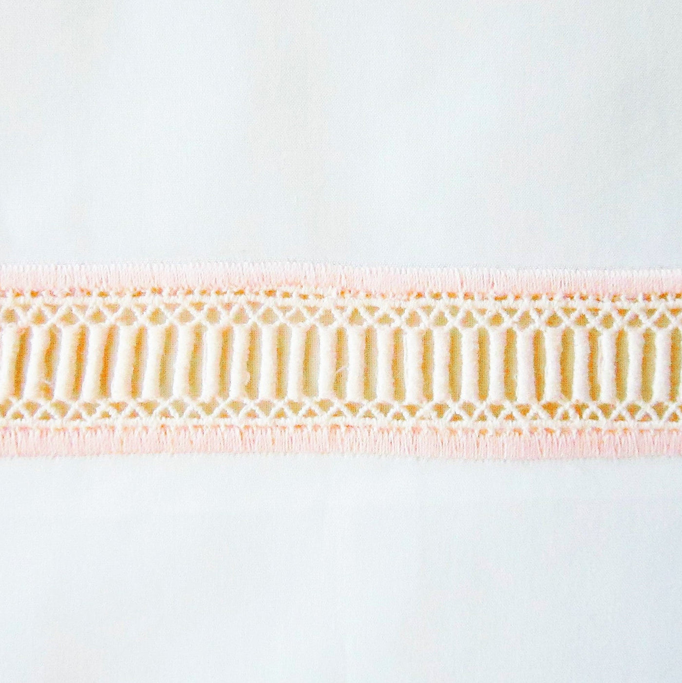 Close-up of Doric blush and gold geometric lace trim on white linen fabric.