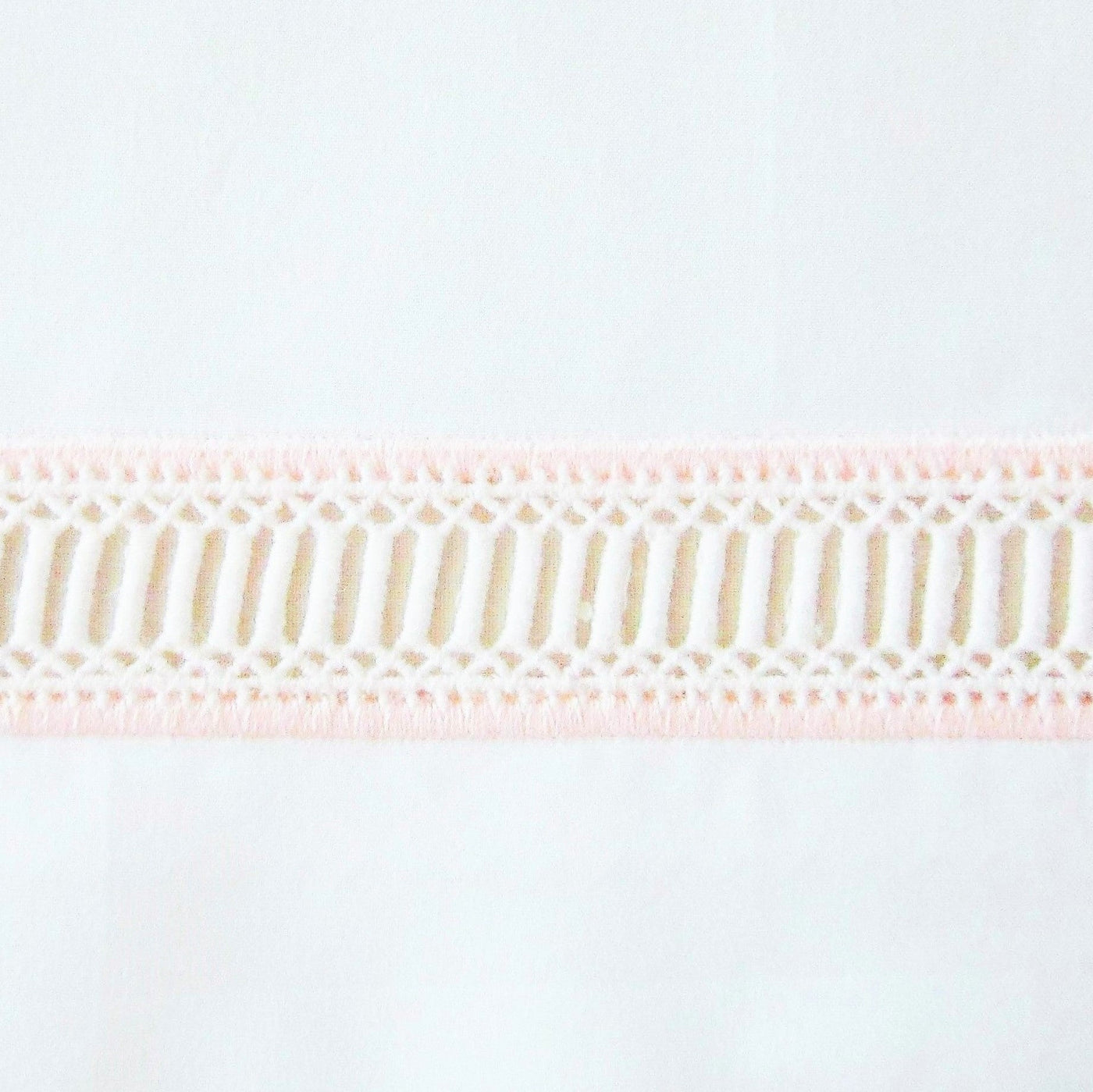 Close-up of blush and ivory geometric lace trim on white cotton fabric.