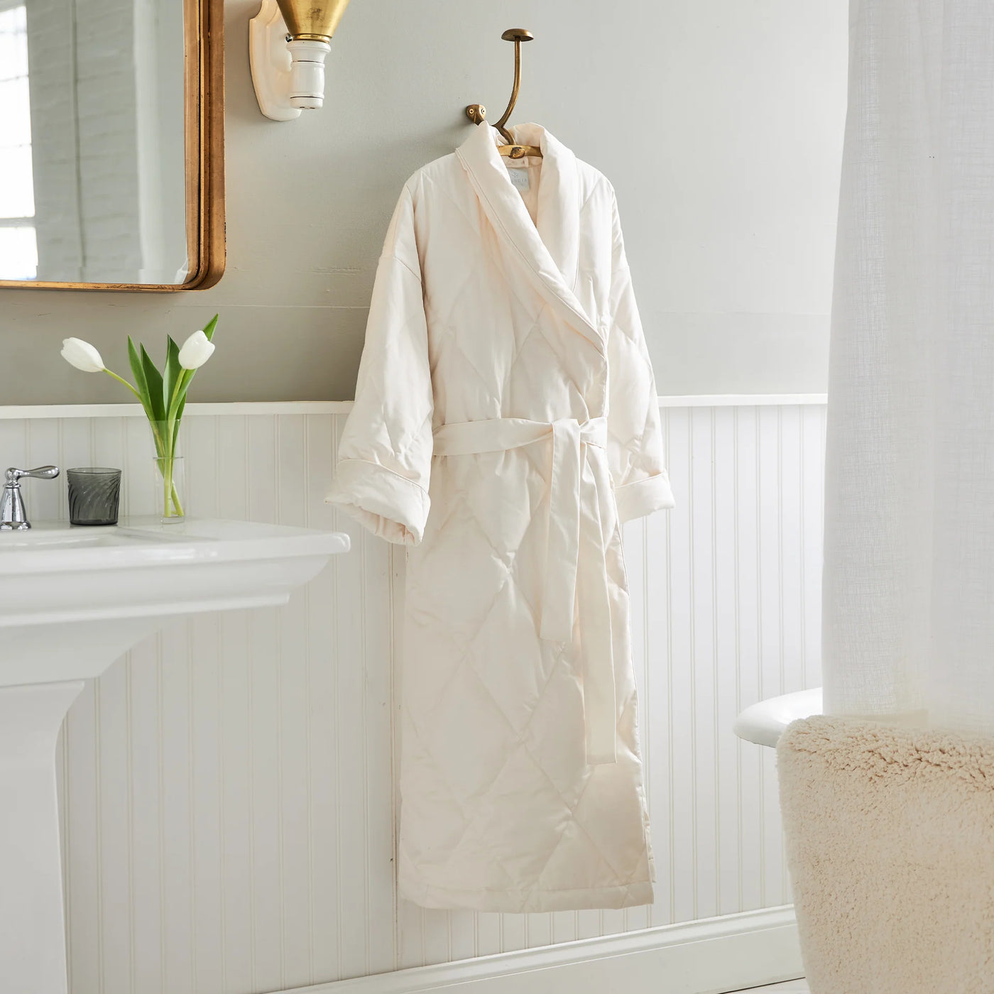 Ivory quilted down robe hanging on brass hook in a white bathroom.