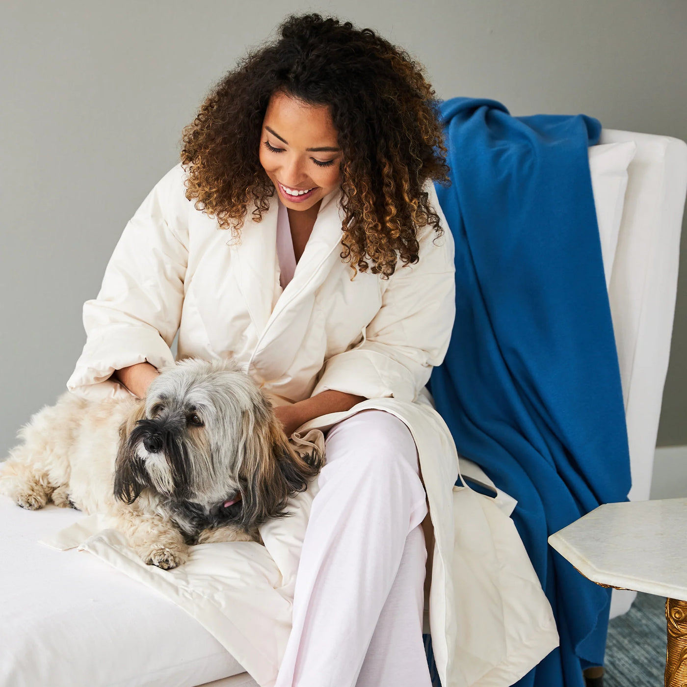 Woman seated on bed petting a dog, wearing ivory Scandia Home down robe over blush pajamas.