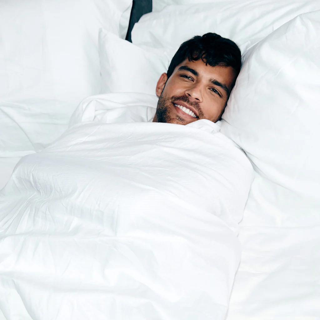 A man lies in bed, smiling, wrapped in the Frette Percale | Duvet Cover by Frette, made of 100% cotton, with his head resting on white pillows.
