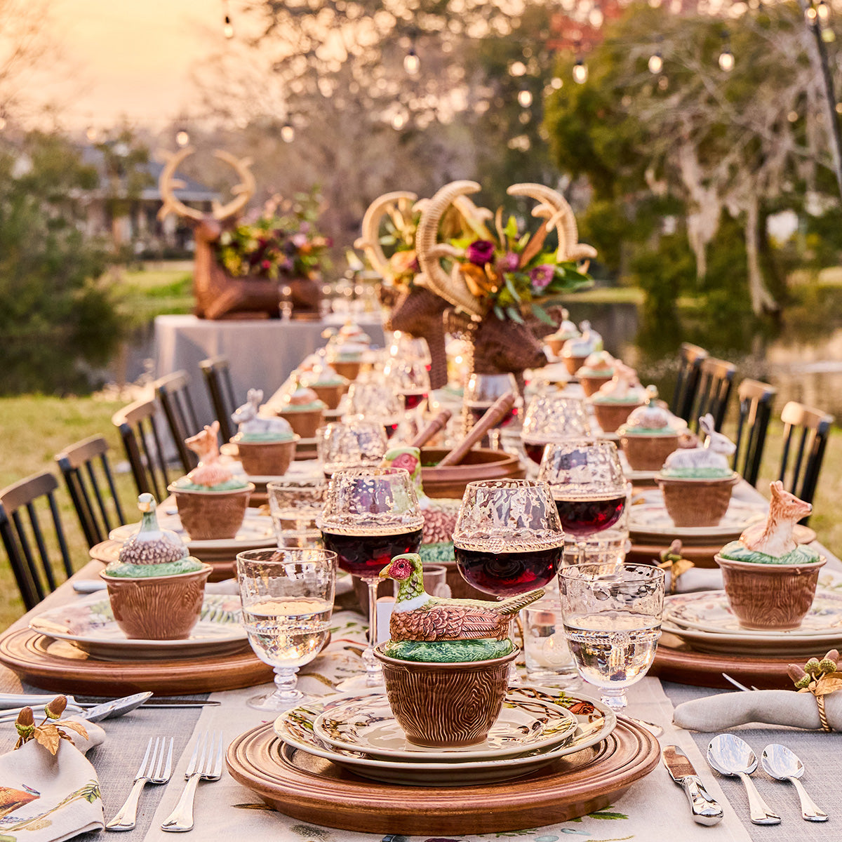 Long outdoor dinner table set with Forest Walk lidded bowls at each place, candles and an antler centerpiece
