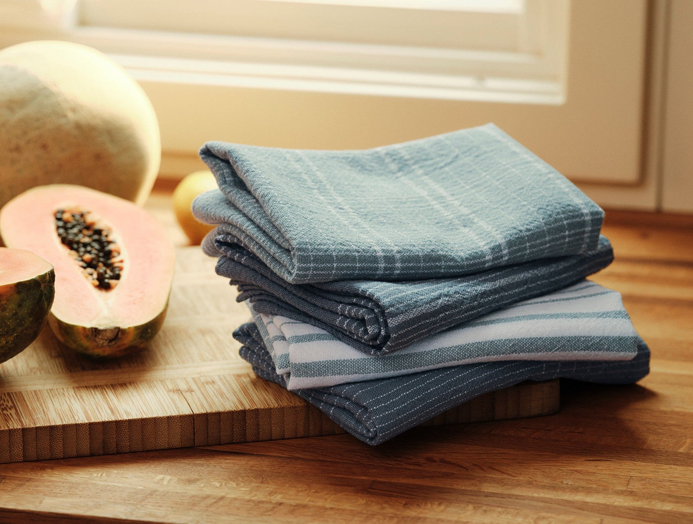 Coyuchi Fillmore kitchen towels stacked on wood cutting board beside sliced papaya, slate blue.