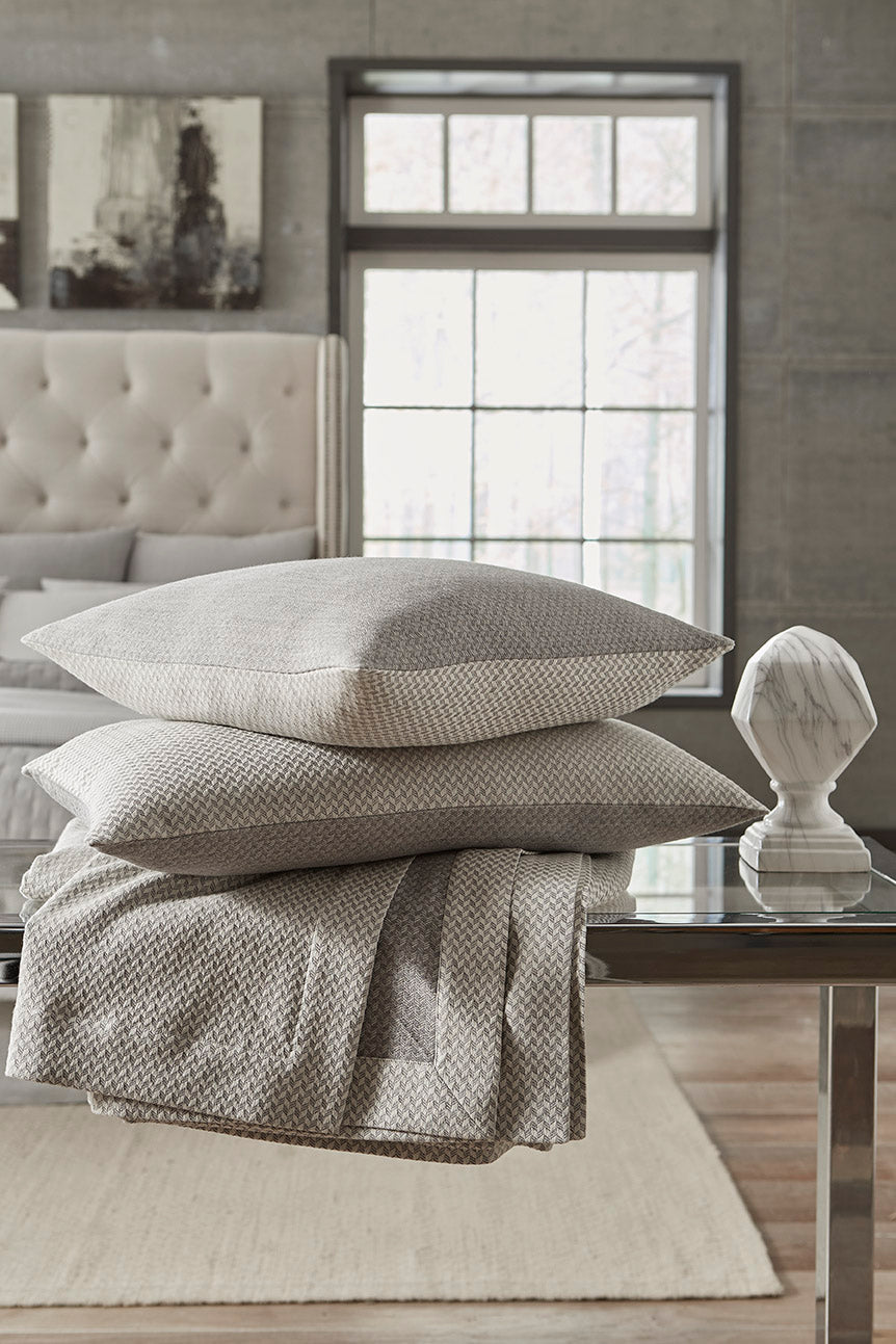 Close-up of stacked Home Treasures Geneva pillows and folded throw in silver-grey woven texture.