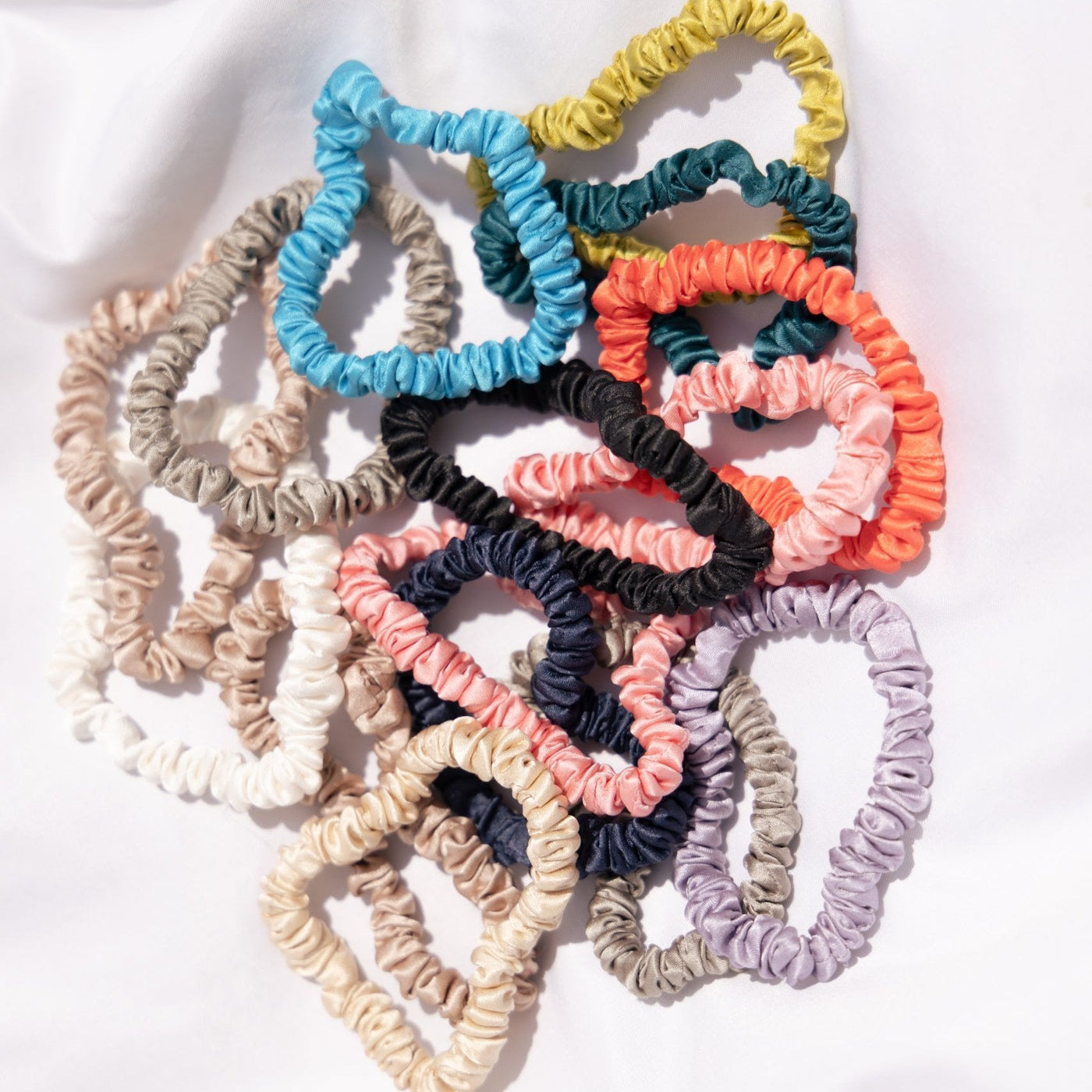 Kumi Kookoon Mulberry Silk Skinny Scrunchies, shown overlapping on white fabric, add a pop of color and help prevent hair breakage.