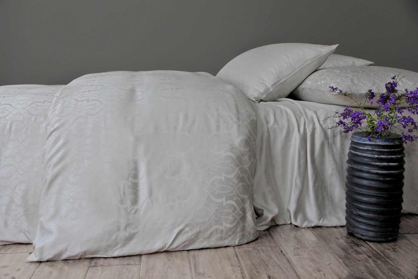Ivory damask-patterned bedding styled on a bed with a dark vase of purple flowers beside it.