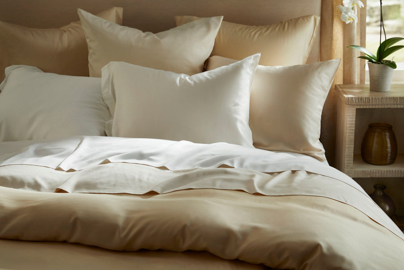 Ivory and champagne Legna Classic bedding styled on a sunlit bed with layered pillows.
