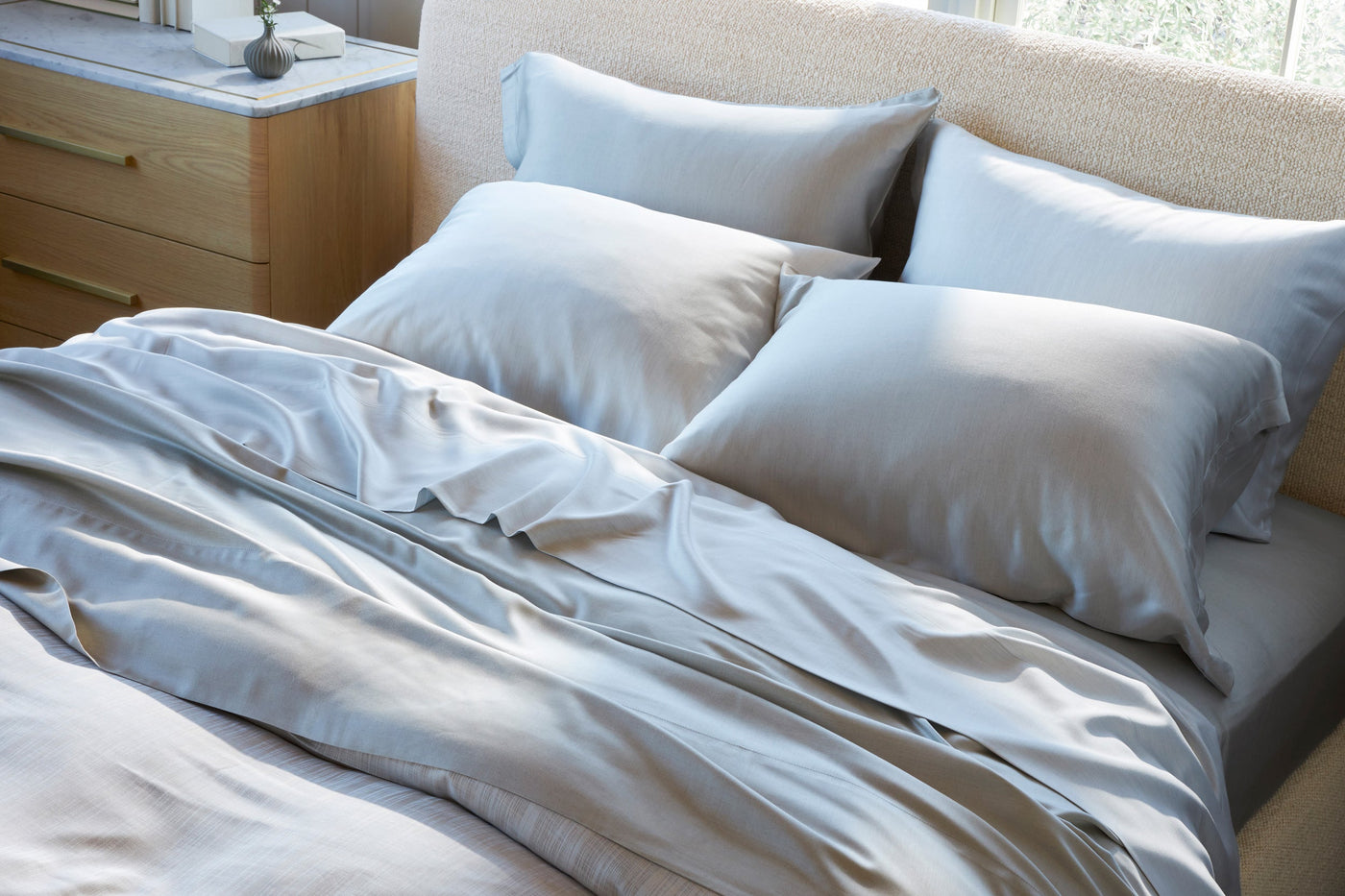 Legna Pisa bedding set in soft blue-grey, draped casually on a sunlit bed with pillows.