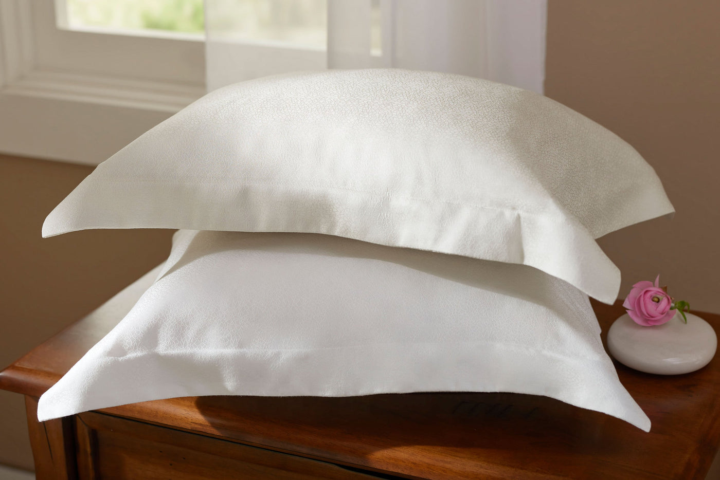 Legna Prieta pillowcases in ivory and white stacked on a wooden dresser beside a pink rose.