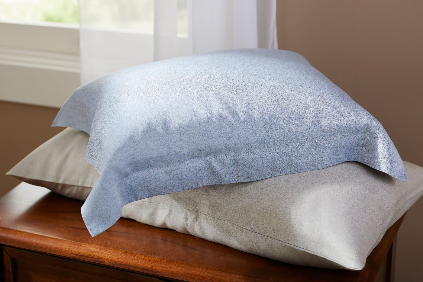 Legna Rimini pillowcases in sky blue and ivory stacked on a wooden surface near a window.