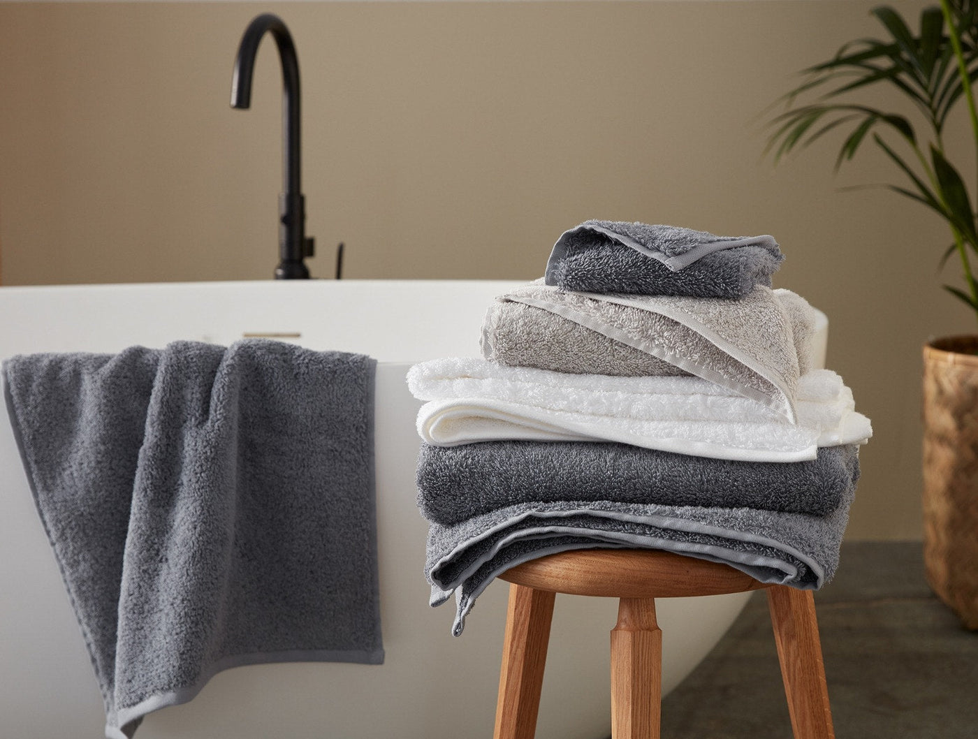 Stack of Coyuchi Cloud Loom towels in white, grey, and charcoal on a stool beside a freestanding bath.