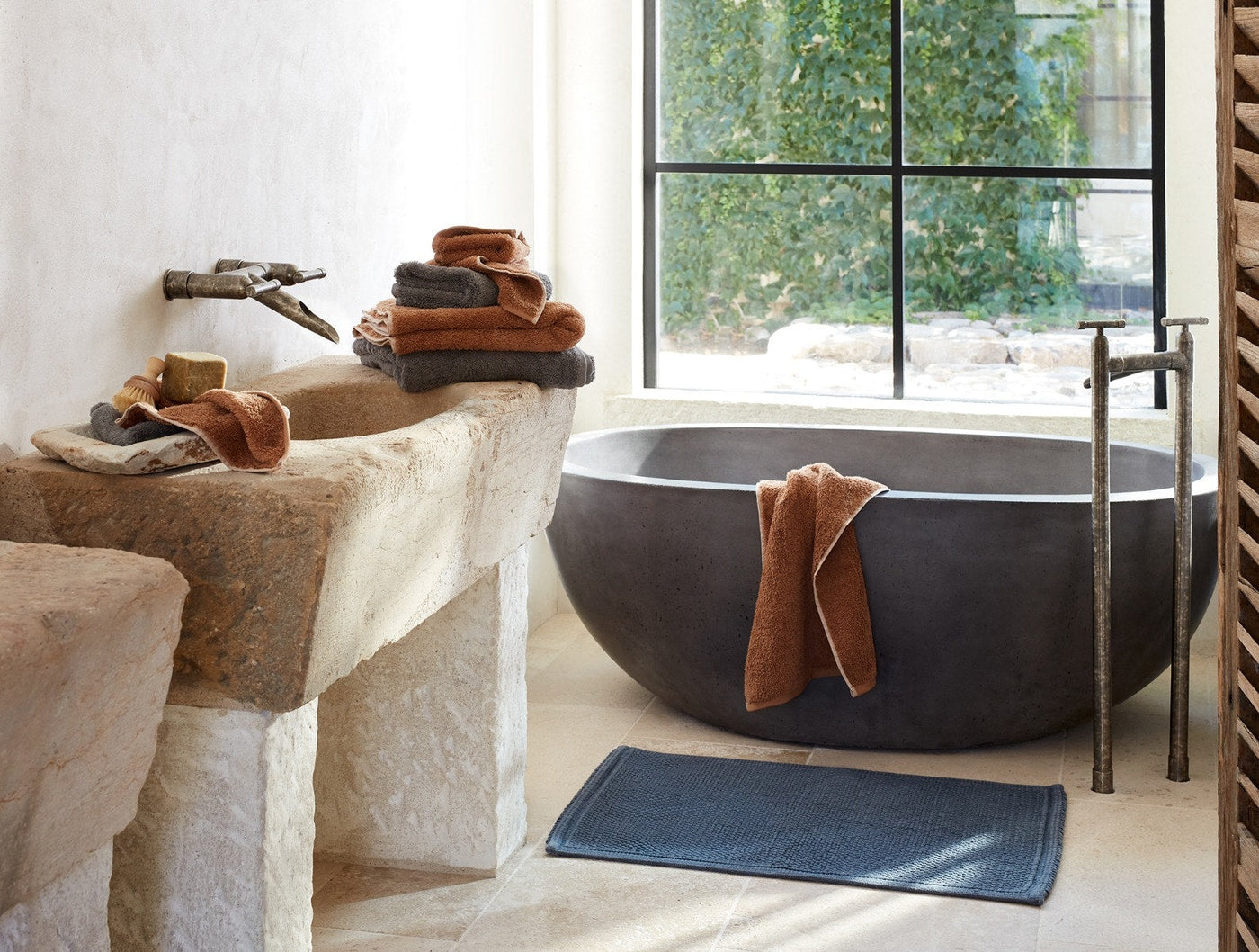 Slate blue bath rug on stone floor beside freestanding tub with rust-toned towels.