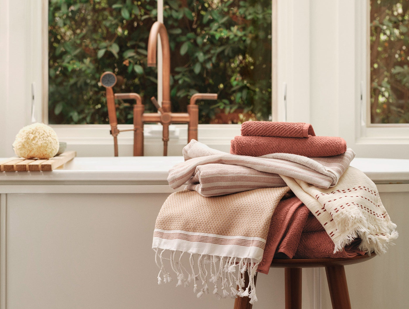 Stacked Coyuchi Mediterranean towel set in warm terracotta and sand tones beside a copper-faucet bathtub.
