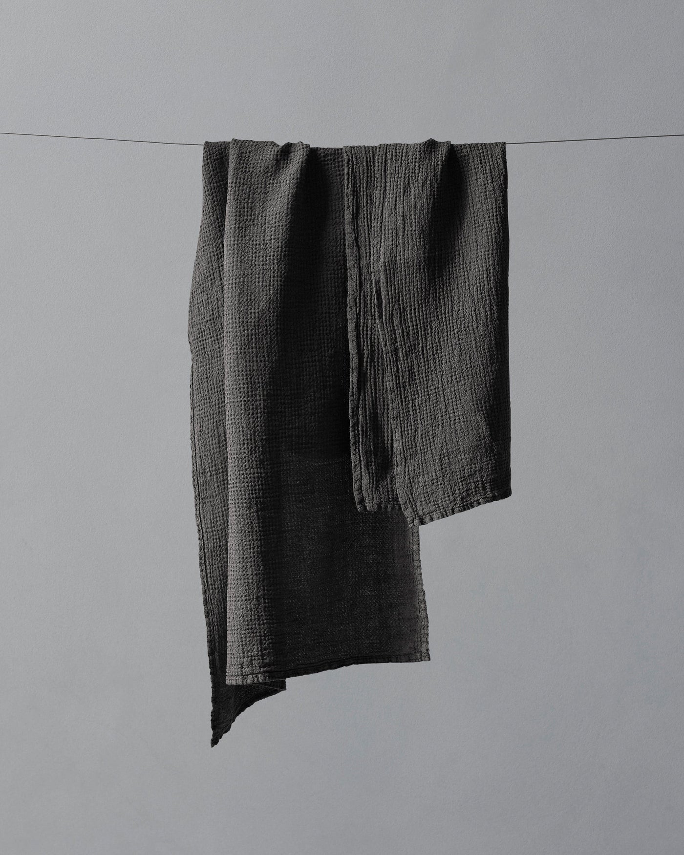 Charcoal textured muslin hand towels draped over a wire against a grey background.