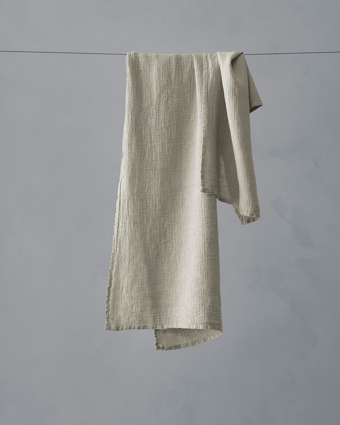 Sage green textured muslin hand towel draped over a washing line against a grey background.