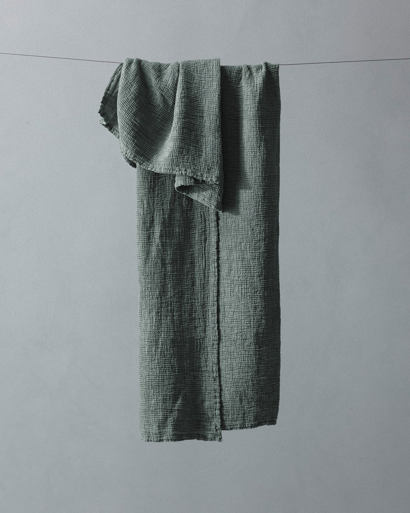 Sage green crinkle-textured cotton towel draped over a thin wire against a grey background.