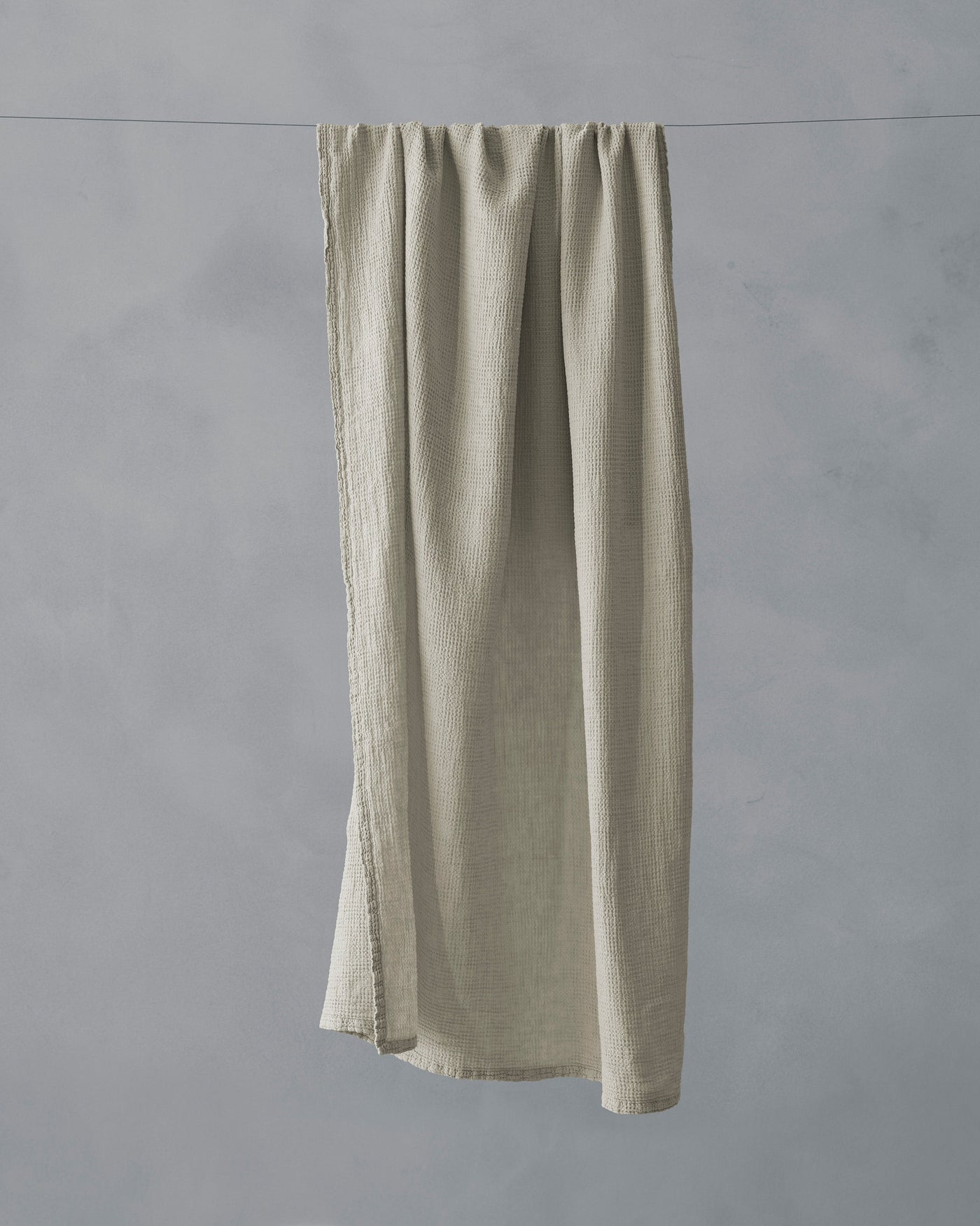 Sage linen bath towel hanging on a line, showing frayed edges and relaxed drape.